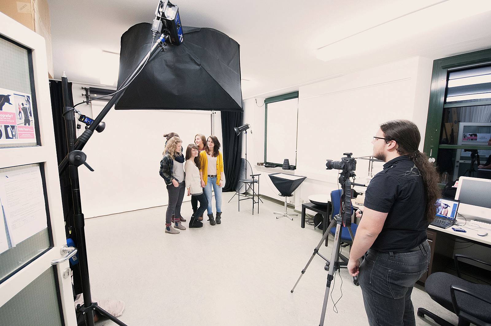 Ein Fotoshooting in einem professionellen, gut ausgestattenen Fotostudio mit verschiedenen Lampen, einer Kameras auf einem Stativen und einem Computer-Tisch. Eine Gruppe von fünf jungen Mädels steht vor einer weißen Leinwand und lässt sich von einem jungen Mann fotografieren.